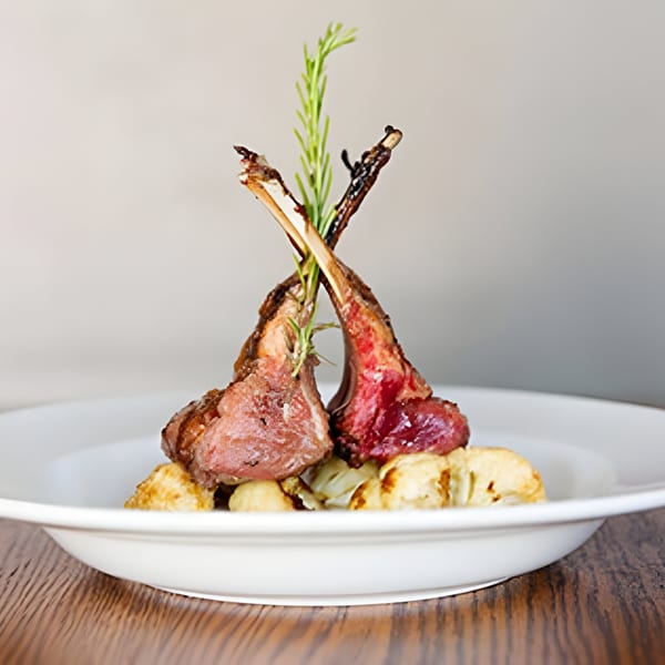 Two lamb chops with rosemary garnish, served on a bed of roasted vegetables in a white bowl.