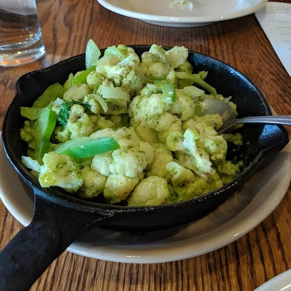 Aro Latin's green cauliflower dish with bell peppers and a spoon in a black cast iron pan.