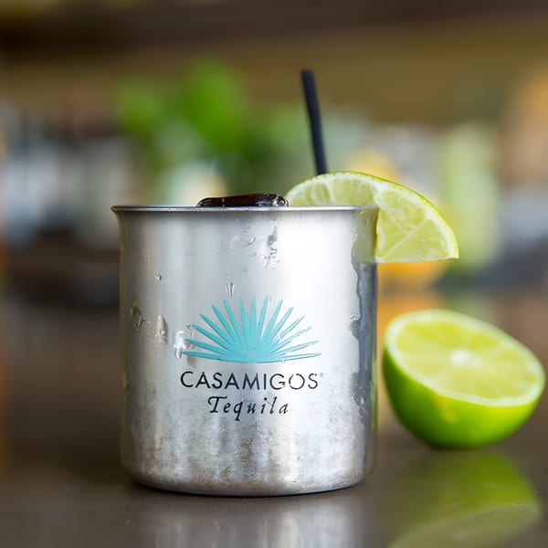 A Casamigos Tequila cocktail in a metal mug with a lime wedge and straw, next to a halved lime.