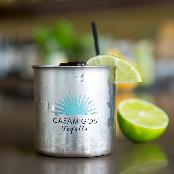 A Casamigos Tequila cocktail in a metal cup with a lime wedge and straw, next to a halved lime.