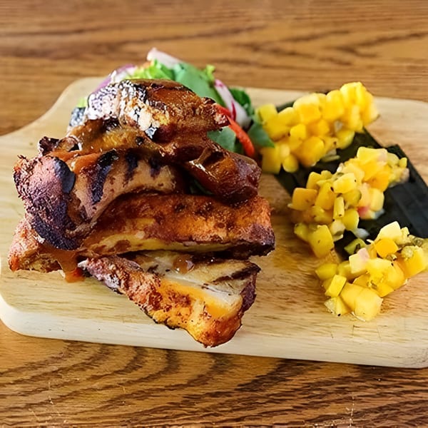 Aro Latin's grilled ribs with sauce, a side salad, and mango salsa on a wooden board.