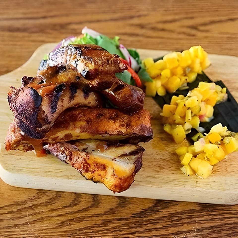 Aro Latin's grilled ribs with sauce, a side salad, and mango salsa on a wooden board.