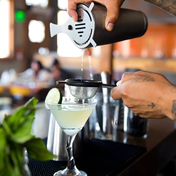 Bartender straining a light green cocktail into a martini glass garnished with a cucumber slice.
