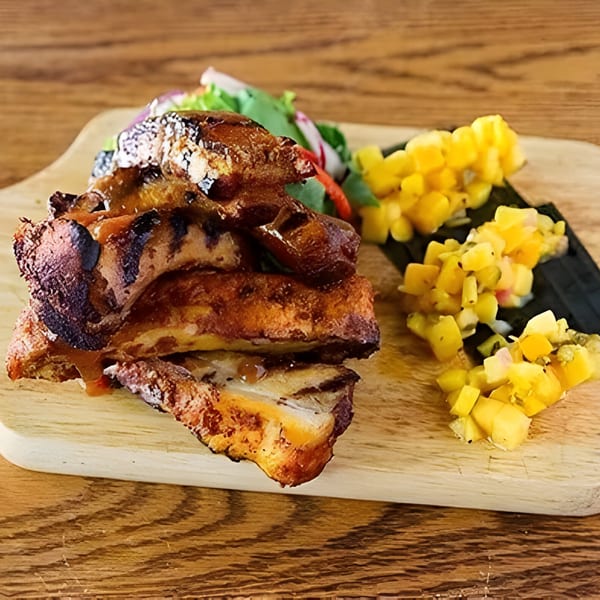 Grilled pork belly slices with mango salsa and a side salad on a wooden cutting board.