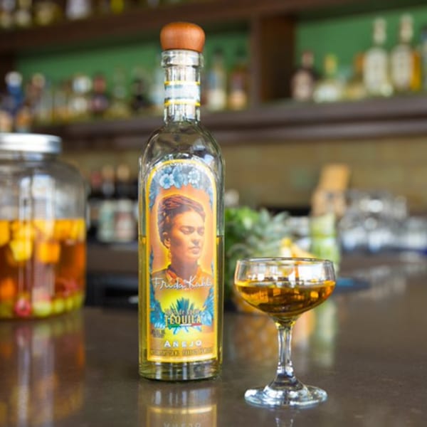 Frida Kahlo Tequila Añejo bottle and a glass of amber liquid on a bar.