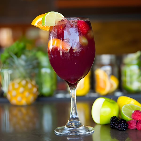 Aro Latin's red sangria in a wine glass, garnished with orange and lime slices, with fruit nearby.